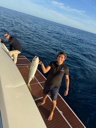 Two smiling men on a boat in the deep blue ocean during an offshore fishing trip — one holding a large catch over the teak swim platform under a clear sunny sky.