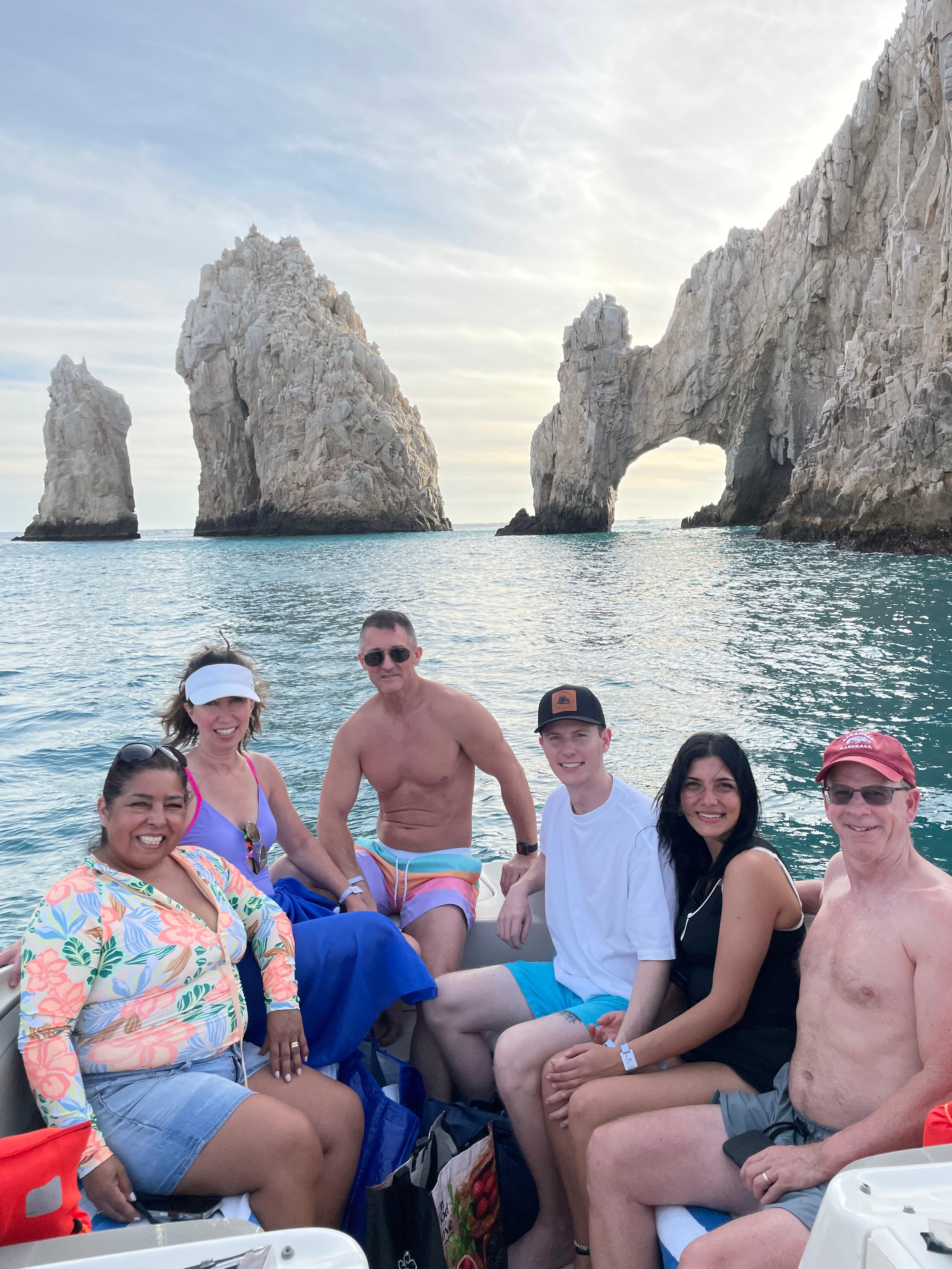 Group of six smiling on a small boat in turquoise water in front of dramatic sea arches and rock formations at Cabo San Lucas during golden hour — fun coastal boat tour scene.