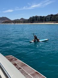 Two people on a paddleboard in clear turquoise water near a sunny beachfront resort; one waves from the board while a boat swim platform is visible in the foreground, with sandy beach, palm trees and hills under a blue sky in the background.