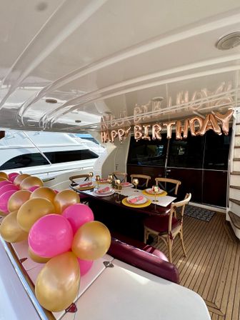 Festive birthday setup on a luxury yacht deck at a marina with rose-gold "HAPPY BIRTHDAY" balloons, pink and gold balloon clusters and an elegant table set with gold-rimmed glassware on a wooden deck.