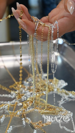 Close-up of a manicured hand holding shimmering gold and silver necklace chains draped over fingers above a tangled jewelry display on a metal tray