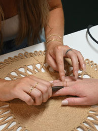 Close-up in a nail salon of a manicurist fitting a delicate gold chain ring on a client's finger over a woven jute mat, pale pink nails and stacked bracelets visible.