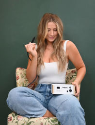 Woman seated cross-legged on a floral armchair, holding a handheld skincare device connected to a control box, wearing a white tank top and jeans against a green backdrop.