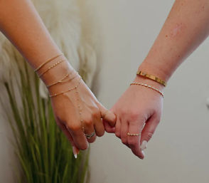 Close-up of two hands linking pinkies in a pinkie-promise, wearing delicate gold bracelets, a hand chain and rings against a neutral indoor background with tall decorative grass.