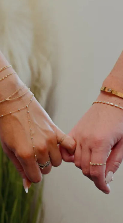 Two hands linking pinkies in a pinky-promise, adorned with gold rings, delicate bracelets and a hand chain, soft indoor plant background — friendship jewelry close-up