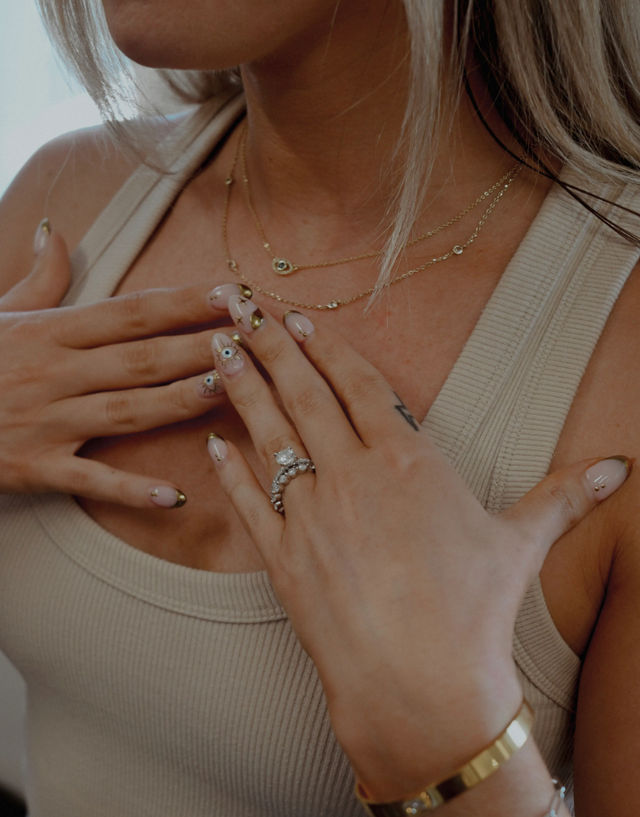 Close-up of a woman’s neckline and hands in a beige ribbed tank, showcasing layered delicate gold necklaces, a diamond ring and gold bracelet, and pink nail art with gold tips and eye motifs.