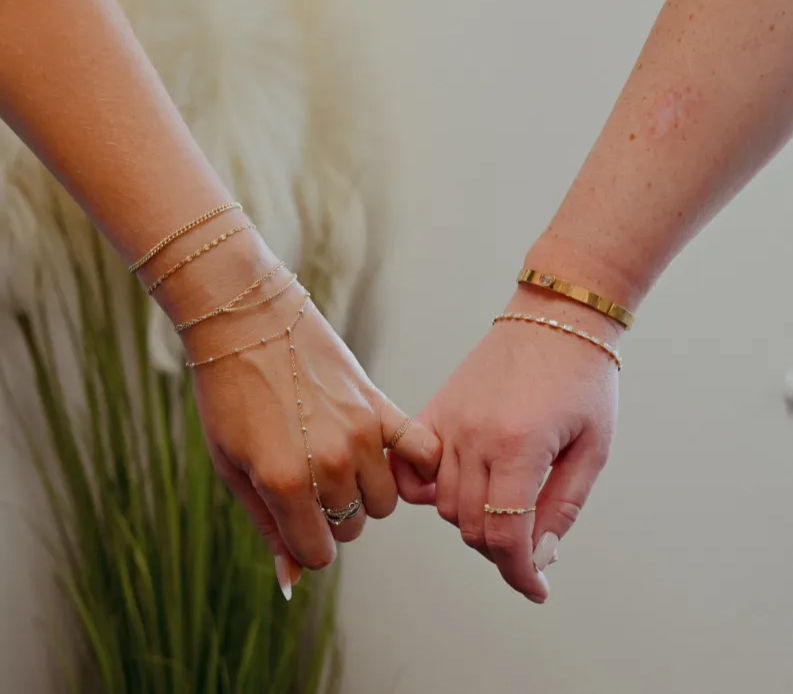 Close-up of two hands linking pinky fingers in a playful promise, wearing delicate gold bracelets, rings and a dainty hand chain against a soft indoor plant background.