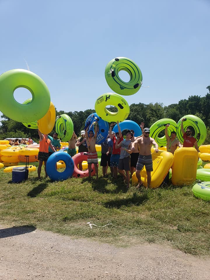 Group of people in swimsuits tossing bright green, blue and yellow inflatable tubes on a sunny grassy riverbank — lively summer tubing outing under a clear blue sky.