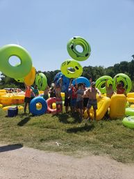 Group of people in swimsuits tossing bright green, blue and yellow inflatable tubes on a sunny grassy riverbank — lively summer tubing outing under a clear blue sky.