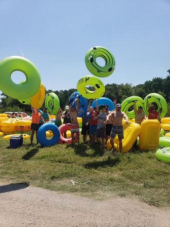 Group of people in swimsuits tossing bright green, blue and yellow inflatable tubes on a sunny grassy riverbank — lively summer tubing outing under a clear blue sky.