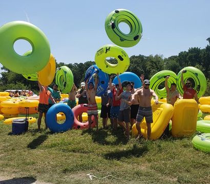 Group of people in swimsuits tossing bright green, blue and yellow inflatable tubes on a sunny grassy riverbank — lively summer tubing outing under a clear blue sky.