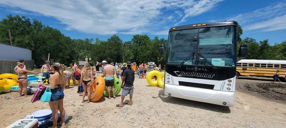 Sun-drenched crowd in swimsuits gathering with colorful inflatable tubes on a sandy river tubing launch next to a parked tour bus and a school bus, trees and blue sky in the background.