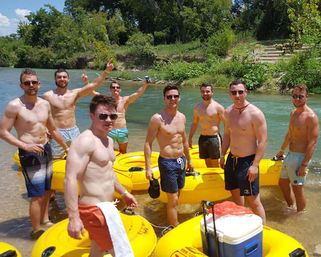 Group of shirtless young men in swim trunks standing in a shallow river next to bright yellow inflatable tubes and a cooler, ready for a sunny outdoor river tubing trip with green trees on the riverbank.