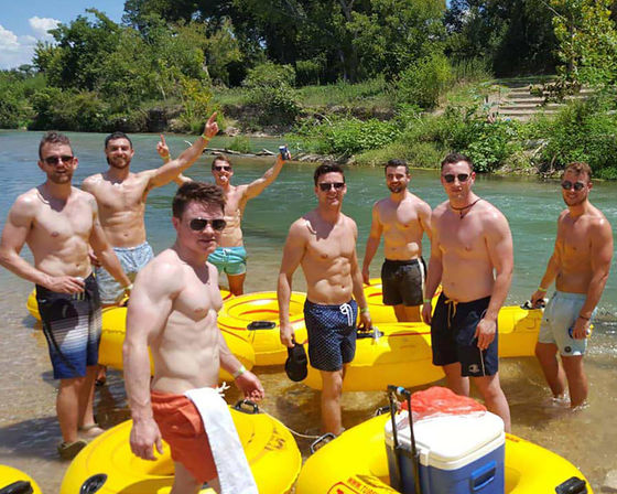 Group of shirtless young men in swim trunks standing in a shallow river next to bright yellow inflatable tubes and a cooler, ready for a sunny outdoor river tubing trip with green trees on the riverbank.