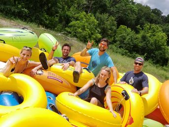 Five smiling adults relaxing on bright yellow inflatable tubes ready for summer river tubing on a sunny, grassy, tree-lined riverbank