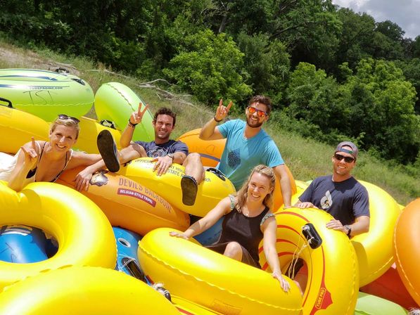 Five smiling adults relaxing on bright yellow inflatable tubes ready for summer river tubing on a sunny, grassy, tree-lined riverbank