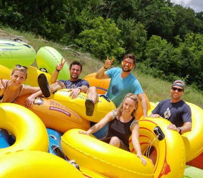 Five smiling adults relaxing on bright yellow inflatable tubes ready for summer river tubing on a sunny, grassy, tree-lined riverbank