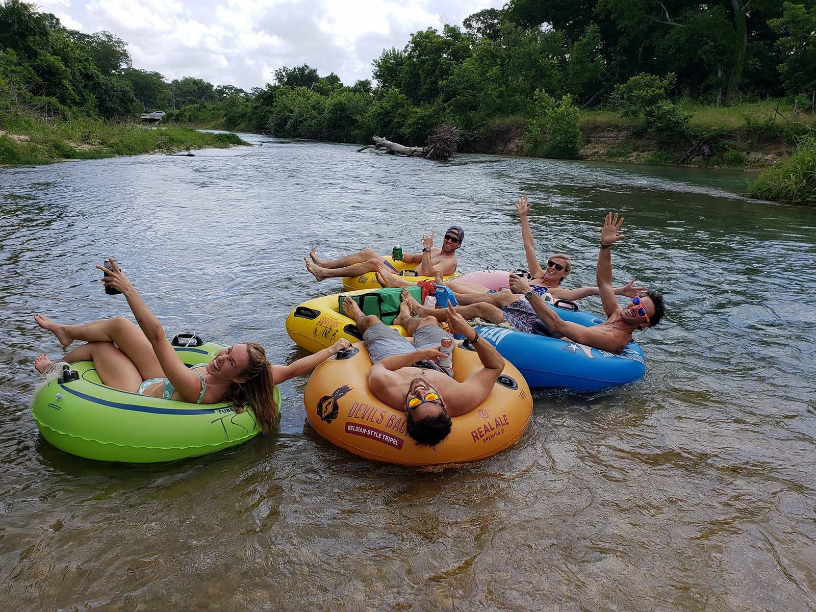 Smiling adults floating on colorful inflatable tubes in a shallow, tree-lined river — summer river tubing and outdoor recreation, hands waving and relaxing on the water.