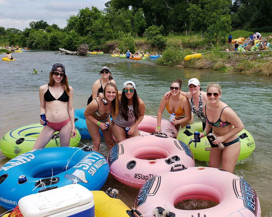 Group of friends on a sunny river tubing outing — people in swimsuits posing on colorful inflatable tubes with canned drinks, grassy riverbank, trees, and other tubers in the background.