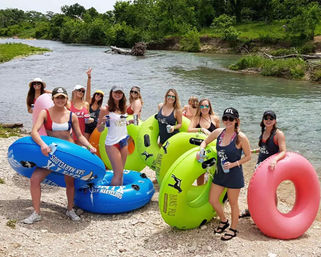 Group of women in swimsuits with colorful inner tubes at a riverbank, holding drinks and smiling before river tubing on a sunny summer day.