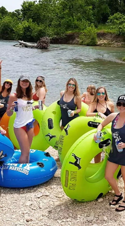 Group of women in swimsuits with colorful inner tubes at a riverbank, holding drinks and smiling before river tubing on a sunny summer day.