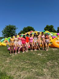 Group of people in colorful swimsuits posing in front of a large pile of bright inflatable tubes on a sunny grassy riverbank, ready for summer tubing under a clear blue sky.