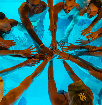 Top-down view of a group of friends in a glowing blue swimming pool, hands stacked in the center for a team cheer
