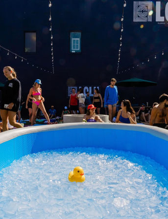 Yellow rubber duck floating in a blue inflatable ice bath at an outdoor urban courtyard ice-bath event with people in swimwear and string lights overhead