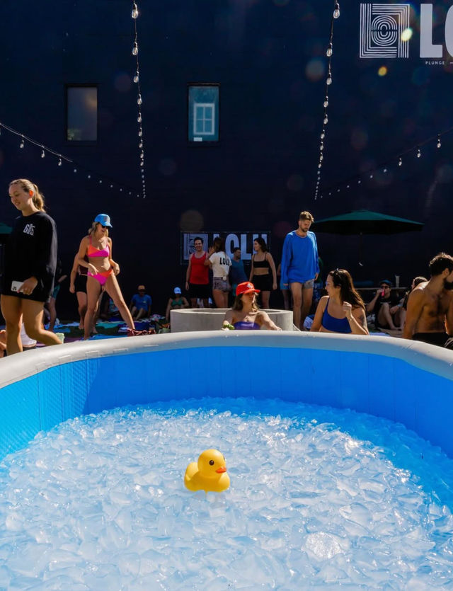 Yellow rubber duck floating in a blue inflatable ice bath at an outdoor urban courtyard ice-bath event with people in swimwear and string lights overhead