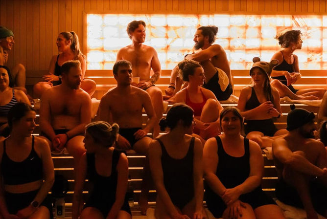 Lively group of adults in swimsuits relaxing and chatting on wooden benches inside a warm-lit spa sauna with a backlit salt wall.