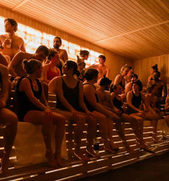 Packed wooden sauna interior with people in swimsuits chatting on tiered benches under warm amber backlit wall — lively spa atmosphere.