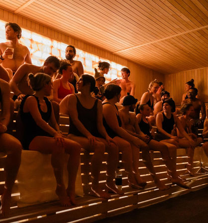 Packed wooden sauna interior with people in swimsuits chatting on tiered benches under warm amber backlit wall — lively spa atmosphere.