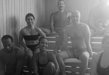 Black-and-white photo of a diverse group of adults in swimwear smiling and relaxing on wooden benches inside an indoor sauna at a spa/wellness center.
