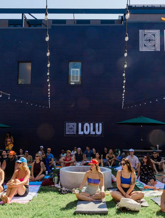 Outdoor yoga and community wellness event in a sunny urban courtyard—participants seated on mats around a circular fire pit under hanging string lights and umbrellas.