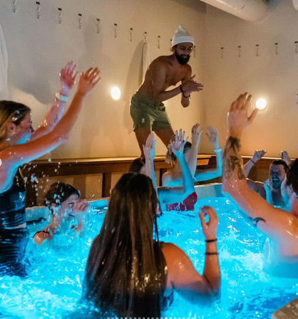 Indoor hot tub party with blue-lit water — people splashing and raising hands while a man in swim shorts and a beanie claps on the tub edge.
