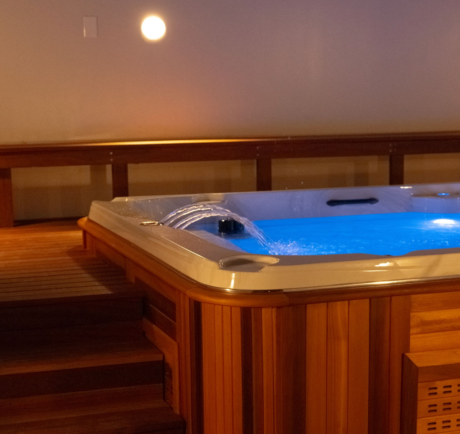 Cozy indoor wooden hot tub on a warm-toned deck, glowing blue water with gentle cascading jets and steps leading up to the spa.