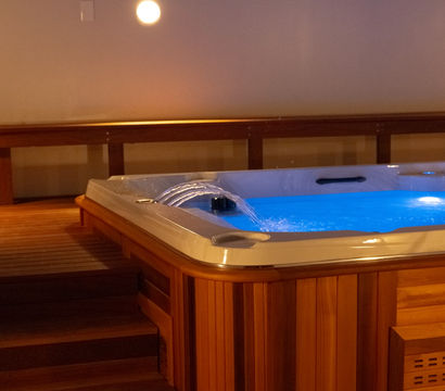 Cozy indoor wooden hot tub on a warm-toned deck, glowing blue water with gentle cascading jets and steps leading up to the spa.