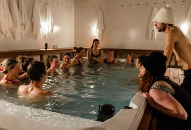 Group of people relaxing and chatting in an indoor hot tub/sauna pool at a wellness spa, some wearing sauna hats with towels hanging on the wall