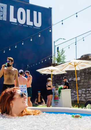 Woman relaxing in an outdoor ice bath filled with crushed ice in a sunny urban courtyard during a summer plunge event, with shirtless attendees, string lights overhead, umbrellas and a vendor booth by a stone wall.