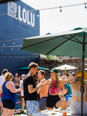 Sunny outdoor patio party with people in swimwear and summer clothes chatting and laughing under green umbrellas and string lights, drinks on tables.