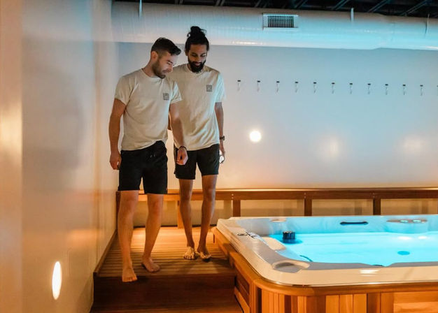 Two men in matching tees and shorts walking barefoot on a wooden deck beside a glowing indoor hot tub with blue water in a modern spa room.