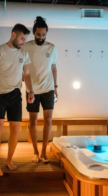 Two men in matching tees and shorts walking barefoot on a wooden deck beside a glowing indoor hot tub with blue water in a modern spa room.