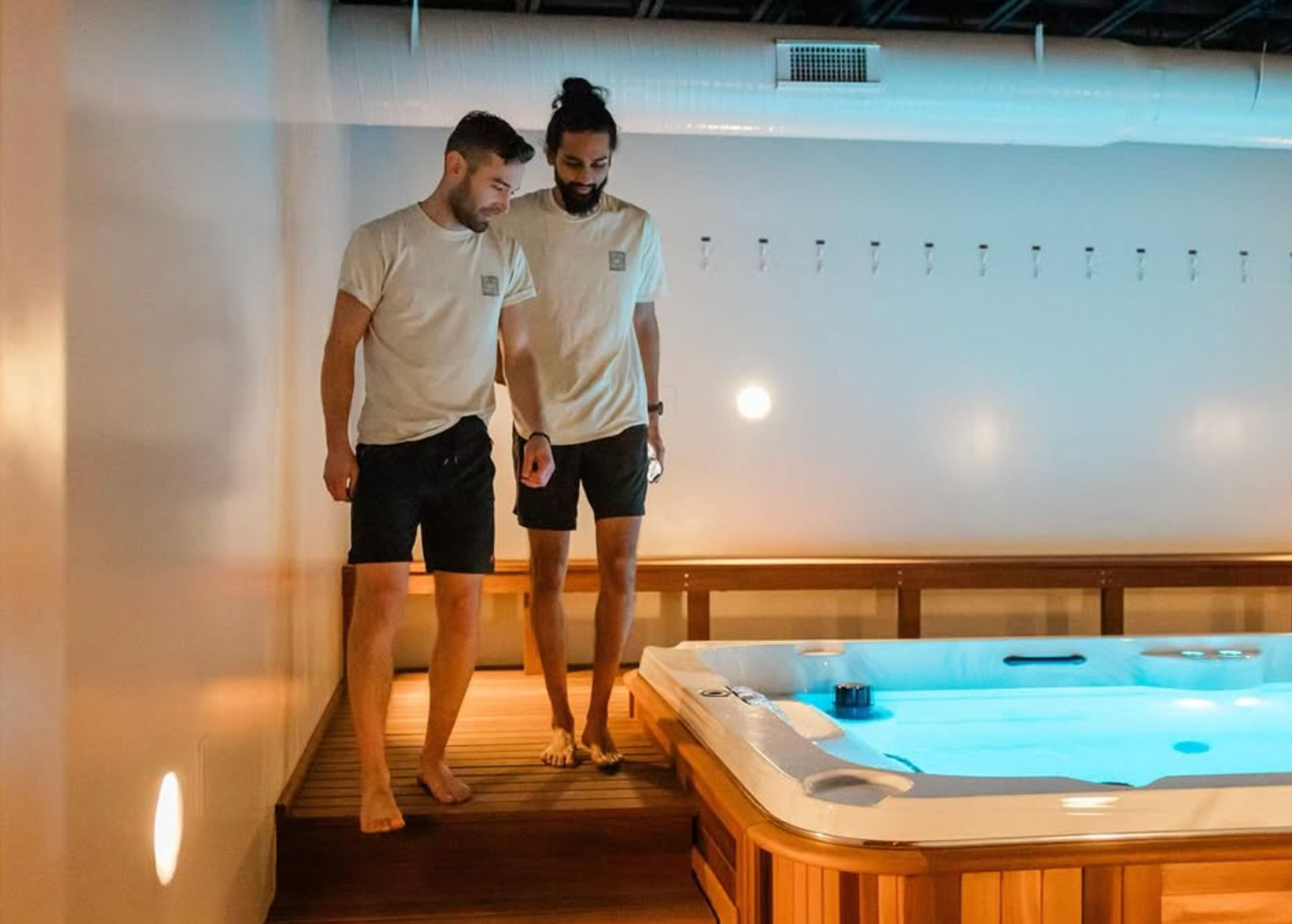Two men in matching tees and shorts walking barefoot on a wooden deck beside a glowing indoor hot tub with blue water in a modern spa room.