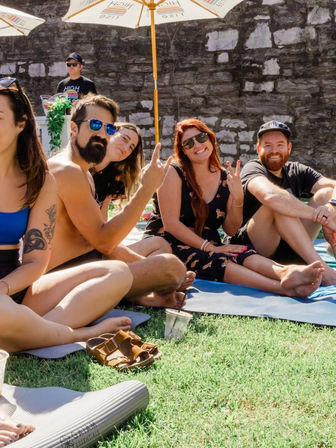 Group of friends lounging on mats on a sunny lawn by a stone wall, wearing sunglasses and making playful hand gestures under umbrellas.