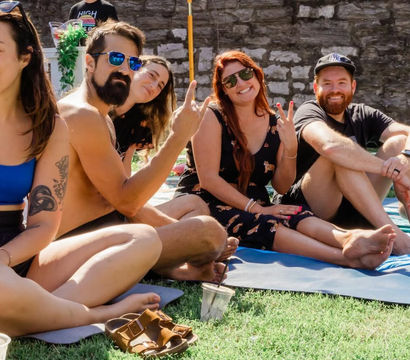 Group of friends lounging on mats on a sunny lawn by a stone wall, wearing sunglasses and making playful hand gestures under umbrellas.