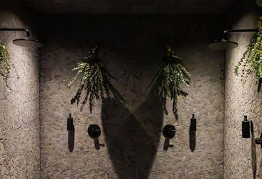 Dimly lit spa shower with three black rain showerheads on a pale mosaic tile wall, hanging ferns above each head and dramatic shadows from soft spotlights