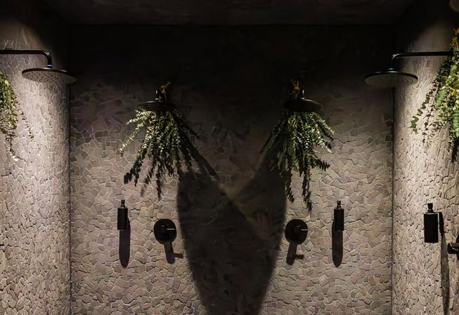 Dimly lit spa shower with three black rain showerheads on a pale mosaic tile wall, hanging ferns above each head and dramatic shadows from soft spotlights