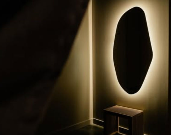 Modern minimalist entryway corner with an organic-shaped backlit wall mirror glowing warm above a small wooden bench, creating moody ambient lighting and sleek interior design vibe.