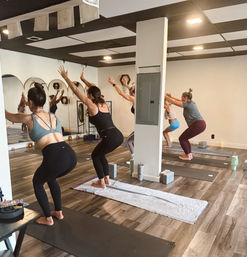 Group yoga class in a bright indoor studio — women on mats practicing chair pose with arms raised, mirrored wall reflection, yoga blocks and water bottles on a wood-look floor.