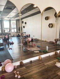 Light-filled mirrored fitness studio with arched gold mirrors and wood floors; a person on a mat doing a glute bridge with a pink exercise ball between knees and holding dumbbells, surrounded by yoga blocks, weights and a water bottle — cozy pilates/strength class vibe.